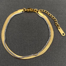 Load image into Gallery viewer, Two-tone layered bracelet featuring a flat 18K gold-plated herringbone chain alongside a delicate silver-tone link chain, with lobster clasp and adjustable extender, displayed on gray fabric.

