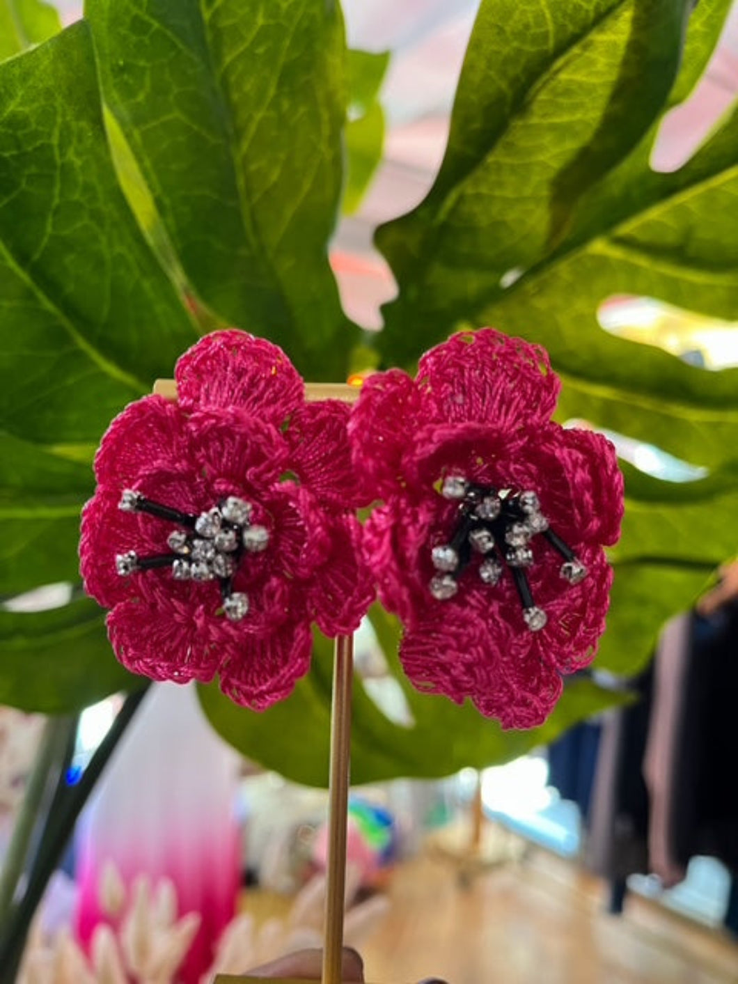 Close-up of fuchsia floral statement earrings on a gold display stand, set against a lush green leaf backdrop. The earrings feature intricate embroidered petals with beaded black and silver crystal centers, capturing a bold and elegant handcrafted design.
