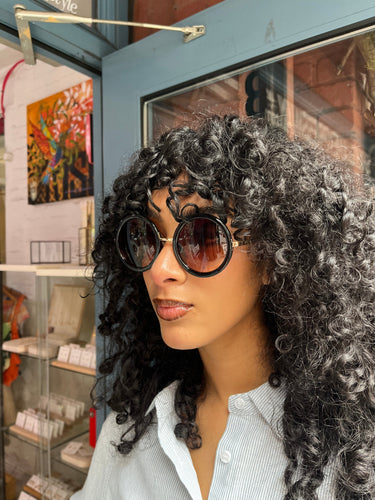 Model wearing oversized round black sunglasses with UVA and UVB protection, standing in front of Glam Expressway Boutique in DUMBO Brooklyn. The sunglasses feature dark gradient lenses and bold frames, styled with a light pinstriped button-down shirt and voluminous curly hair. A fashionable summer accessory perfect for city chic and sun-safe style.
