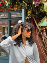 Load image into Gallery viewer, Model wearing oversized round black sunglasses with UVA and UVB protection, standing in front of Glam Expressway Boutique in DUMBO Brooklyn. The sunglasses feature dark gradient lenses and bold frames, styled with a light pinstriped button-down shirt and voluminous curly hair. A fashionable summer accessory perfect for city chic and sun-safe style.
