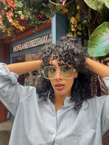 Model wearing oversized square green sunglasses with transparent frames and tinted lenses, posing in front of Glam Expressway Boutique in DUMBO, Brooklyn. Trendy summer eyewear offering UV protection and bold retro style.