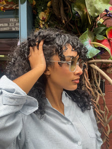 Model wearing oversized square green sunglasses with transparent frames and tinted lenses, posing in front of Glam Expressway Boutique in DUMBO, Brooklyn. Trendy summer eyewear offering UV protection and bold retro style.