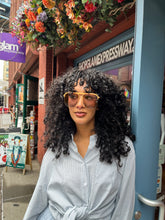Load image into Gallery viewer, Model wearing oversized rose gold aviator sunglasses with peach-toned frames and tinted lenses, standing outside Glam Expressway boutique in DUMBO, Brooklyn, surrounded by vibrant storefront signage and flowers.
