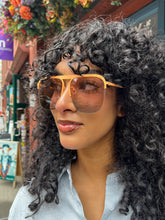 Load image into Gallery viewer, Model wearing oversized rose gold aviator sunglasses with peach-toned frames and tinted lenses, standing outside Glam Expressway boutique in DUMBO, Brooklyn, surrounded by vibrant storefront signage and flowers.
