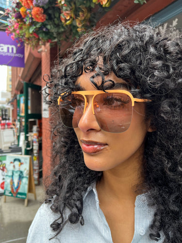 Model wearing oversized rose gold aviator sunglasses with peach-toned frames and tinted lenses, standing outside Glam Expressway boutique in DUMBO, Brooklyn, surrounded by vibrant storefront signage and flowers.