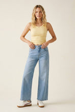 Load image into Gallery viewer, A woman modeling a pair of light-wash wide-leg blue jeans with a pale yellow ribbed tank top and white sneakers, standing against a solid light beige studio background.

