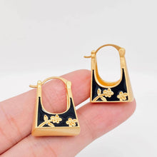 Load image into Gallery viewer, Black enamel handbag-shaped huggie earrings with gold-tone trim and delicate carved floral details.
