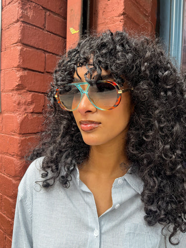 Model wearing oversized colorblock gradient sunglasses with orange and teal frames, gold accents, and tinted lenses, posing outside Glam Expressway boutique in DUMBO, Brooklyn, with signature red brick wall and window storefront in the background.