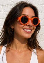 Load image into Gallery viewer, Woman wearing orange sunglasses against a white brick wall
