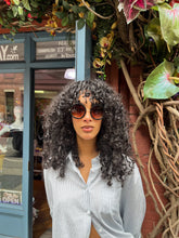 Load image into Gallery viewer, Model wearing oversized round black sunglasses with UVA and UVB protection, standing in front of Glam Expressway Boutique in DUMBO Brooklyn. The sunglasses feature dark gradient lenses and bold frames, styled with a light pinstriped button-down shirt and voluminous curly hair. A fashionable summer accessory perfect for city chic and sun-safe style.
