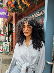 Model wearing oversized rose gold aviator sunglasses with peach-toned frames and tinted lenses, standing outside Glam Expressway boutique in DUMBO, Brooklyn, surrounded by vibrant storefront signage and flowers.