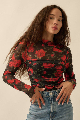 Woman wearing a black long-sleeve top with red floral pattern and blue jeans against a plain background