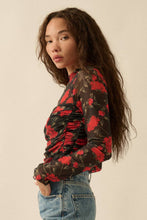 Load image into Gallery viewer, Woman wearing a floral blouse with red and black flowers on a beige background
