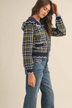 Load image into Gallery viewer, Model wearing a ruffled plaid knit cardigan with puff sleeves, scalloped trim, button-front V neckline, and contrast cable-knit yoke, styled with denim jeans.
