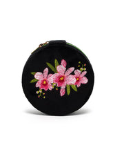 Load image into Gallery viewer, Black velvet round travel jewelry case with embroidered pink orchids, green leaves and contrasting green zipper.
