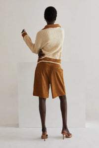 Model wearing high-waisted brown corduroy Bermuda shorts paired with a loose, deep brown blouse and nude heeled mules. She stands confidently next to a wooden stool against a minimal studio backdrop, highlighting the timeless, year-round style of the shorts.