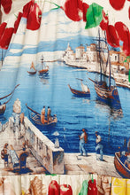 Load image into Gallery viewer, Model wearing a cream halter maxi dress featuring colorful Mediterranean coastal scenes, cherries, lemons, and postcard-inspired prints with a flowing tiered silhouette.

