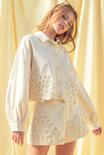 Load image into Gallery viewer, Model wearing an écru cropped shirt jacket with oversized floral appliqué embroidery and matching neutral tailored shorts in a soft resort-inspired setting.
