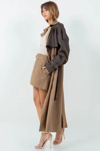 A long brown trench coat featuring a mix of vegan leather and traditional trench materials, offering a unique and chic style. 