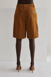 Model wearing high-waisted brown corduroy Bermuda shorts paired with a loose, deep brown blouse and nude heeled mules. She stands confidently next to a wooden stool against a minimal studio backdrop, highlighting the timeless, year-round style of the shorts.