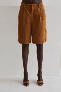 Model wearing high-waisted brown corduroy Bermuda shorts paired with a loose, deep brown blouse and nude heeled mules. She stands confidently next to a wooden stool against a minimal studio backdrop, highlighting the timeless, year-round style of the shorts.