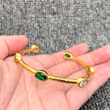 Load image into Gallery viewer, 18K gold-plated open cuff bangle with a hammered textured finish and oval emerald-green and clear zircon stones in gold settings, displayed in the palm of a hand.

