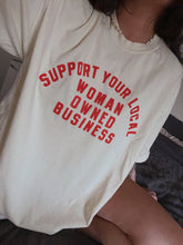 Load image into Gallery viewer, Cream 100% cotton graphic T-shirt with bold red lettering reading "Support Your Local Woman-Owned Business," shown on a model wearing the relaxed-fit tee.
