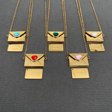 Load image into Gallery viewer, Five gold-tone necklace pendants displayed on a dark gray textured background. Each necklace features an envelope-shaped charm with a different colored heart gemstone (blue, green, white, red, pink) and a small rectangular tag engraved with 'Love You'.
