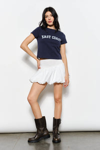 Woman wearing a navy blue 'East Coast' t-shirt, white skirt, and black boots on a white background
