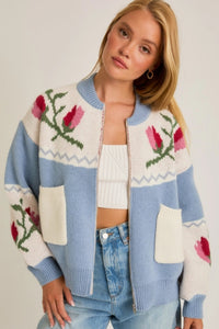 A light blue and cream color-block cardigan sweater featuring a vibrant floral intarsia knit design, seamlessly blending the comfort of a sweater with the structure of a cardigan jacket for effortless style. 