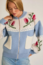 Load image into Gallery viewer, A light blue and cream color-block cardigan sweater featuring a vibrant floral intarsia knit design, seamlessly blending the comfort of a sweater with the structure of a cardigan jacket for effortless style. 
